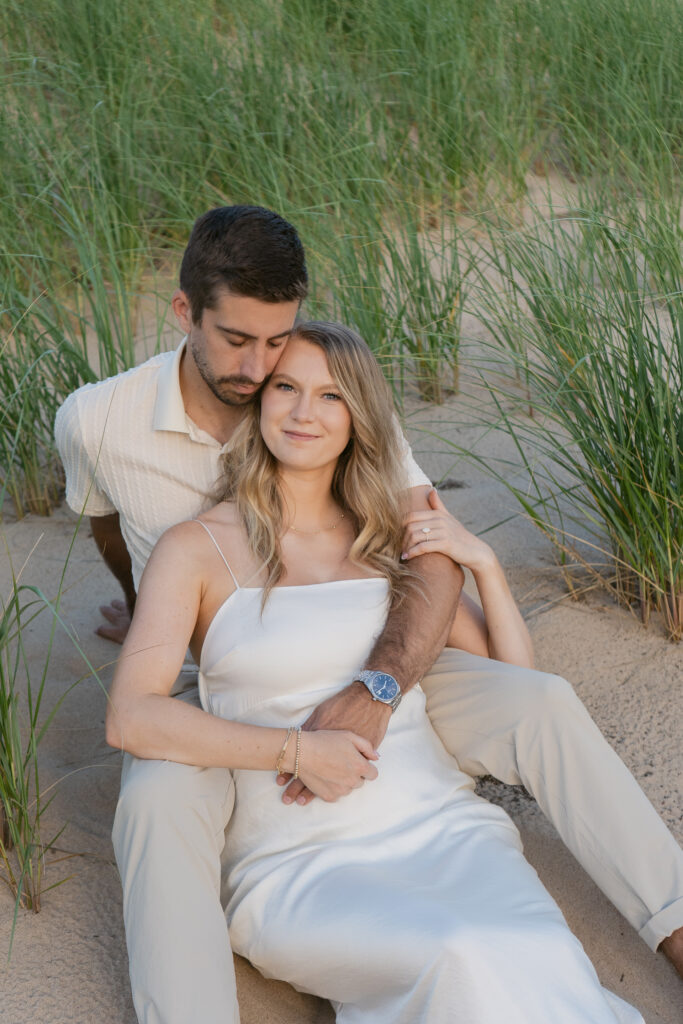 Image of couple sitting together in the sand on the grassy dunes of Oval Beach in Michigan during golden hour engagement session.