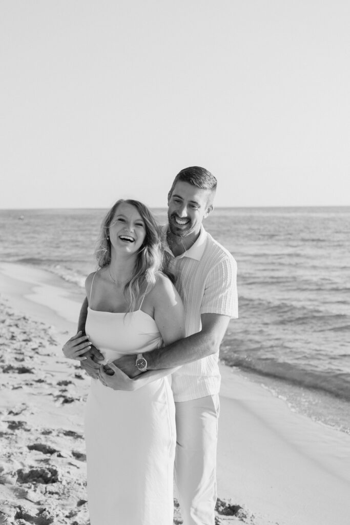Black and white image of couple embracing and laughing on the shore at Oval Beach in Michigan during golden hour engagement session.