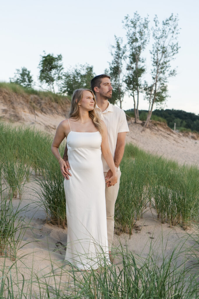Image of couple standing together on the grassy dunes of Oval Beach in Michigan at golden hour looking out at the water.